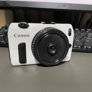 May include: A white Canon EOS camera with a black lens. The camera has a black rubber grip on the right side.