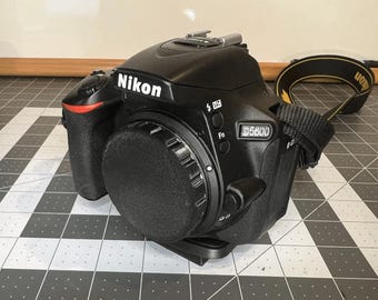May include: A black Nikon D5600 digital SLR camera with a black lens cap attached. The camera has a black strap with yellow stitching.