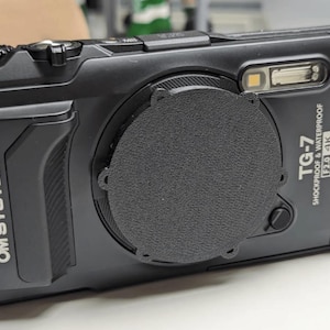 Puede incluir: Cámara digital negra OM System TG-7 a prueba de golpes y resistente al agua. La cámara tiene una tapa de lente, un flash y el texto "TG-7 SHOCKPROOF & WATERPROOF F2.0 4K". La cámara está sobre una superficie blanca.