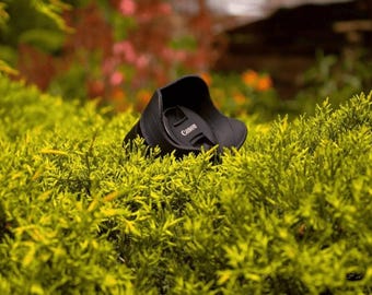 May include: A black Canon lens cap is partially hidden in a bed of green foliage.