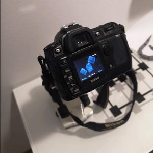 May include: A black Nikon DSLR camera with a blue screen displaying a photo of blue ice cubes. The camera is mounted on a tripod.