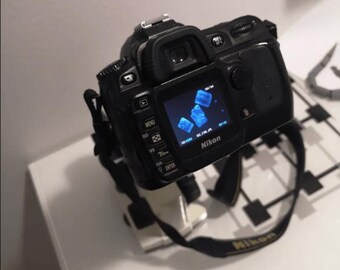 May include: A black Nikon DSLR camera with a blue screen displaying a photo of blue ice cubes. The camera is mounted on a tripod.
