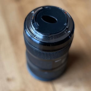 May include: A black camera lens with a ridged body and a circular opening at the top. The lens has a metallic ring and several small markings. The lens is designed for use with a camera.