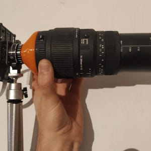 May include: A black camera lens with a 70-300mm zoom range and a 4-5.6 aperture setting. The lens is attached to a tripod and has an orange plastic adapter.