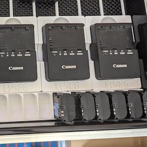 May include: Three black Canon LP-E6 battery chargers with a row of six black LP-E6 batteries below them.