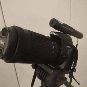 Könnte beinhalten: Eine schwarze Nikon D5300 Kamera mit einem schwarzen Objektiv, das an einem Stativ befestigt ist. Die Kamera hat ein schwarzes externes Mikrofon, das oben angebracht ist.