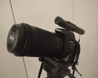 May include: A black Nikon D5300 camera with a black lens attached to a tripod. The camera has a black external microphone attached to the top.