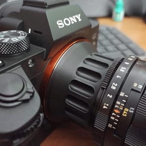 Op de afbeelding: Een zwarte Sony camera met een zwarte lens bevestigd. De lens heeft een zilveren ring met markeringen voor diafragma en afstand in voet en meter. De camera heeft een zwarte draaiknop met het woord "AUTO" erop.