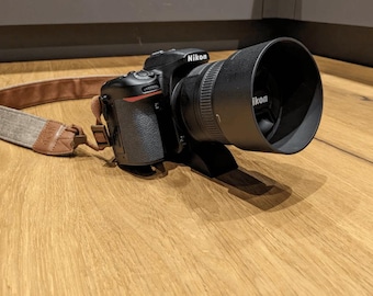 May include: A black Nikon DSLR camera with a lens and a lens hood. The camera is attached to a brown and grey camera strap.