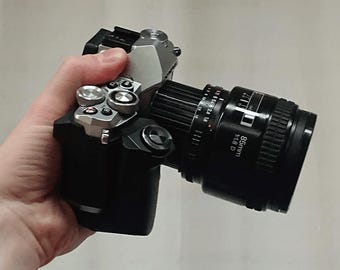 May include: A silver and black camera with an 85mm f1.8 lens attached. The camera has multiple buttons and dials.