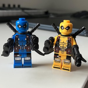 May include: Two Lego minifigures of Deadpool, one in blue and one in yellow, both with black guns and swords.