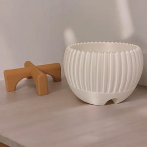 Pode incluir: Um vaso de cer&acirc;mica branco, canelado, com uma borda ondulada, est&aacute; ao lado de um pequeno suporte de madeira castanha para plantas. O vaso tem formato arredondado e superf&iacute;cie texturizada. O suporte tem um design arqueado.