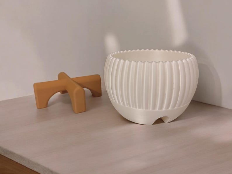 Pode incluir: Um vaso de cer&acirc;mica branco, canelado, com uma borda ondulada, est&aacute; ao lado de um pequeno suporte de madeira castanha para plantas. O vaso tem formato arredondado e superf&iacute;cie texturizada. O suporte tem um design arqueado.