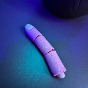 May include: A segmented, light purple finger model with a light blue fingernail. The finger is broken into three sections, with visible wood grain texture. The model is resting on a dark blue surface.