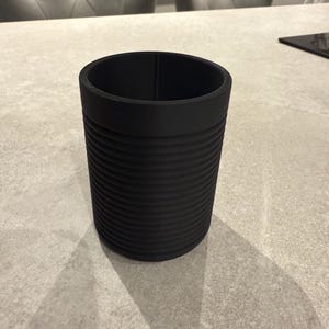 Pode incluir: Um recipiente cil&iacute;ndrico preto com um exterior canelado. O recipiente &eacute; feito de um material fosco e tem um topo liso e aberto. O recipiente est&aacute; sobre uma superf&iacute;cie cinza claro.