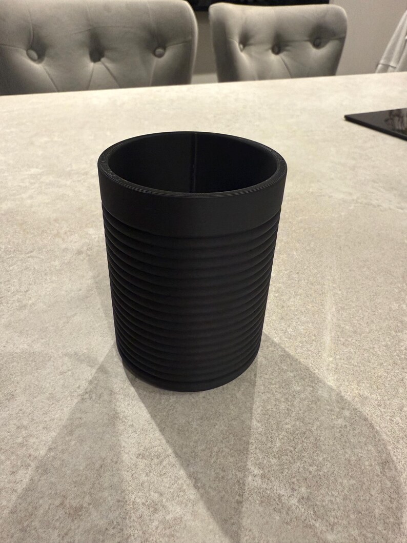 Pode incluir: Um recipiente cil&iacute;ndrico preto com um exterior canelado. O recipiente &eacute; feito de um material fosco e tem um topo liso e aberto. O recipiente est&aacute; sobre uma superf&iacute;cie cinza claro.