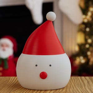 May include: A festive Christmas decoration shaped like Santa Claus. The figure has a white body, a red hat with a white ball on top, and a red nose. The background includes a blurred image of a Christmas tree and a Santa Claus figure.