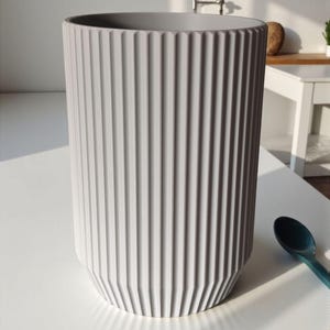 May include: A tall, gray cylindrical vase with vertical ridged detailing. The vase has a slightly tapered base and a smooth, open top. A teal spoon rests on the white surface next to the vase.