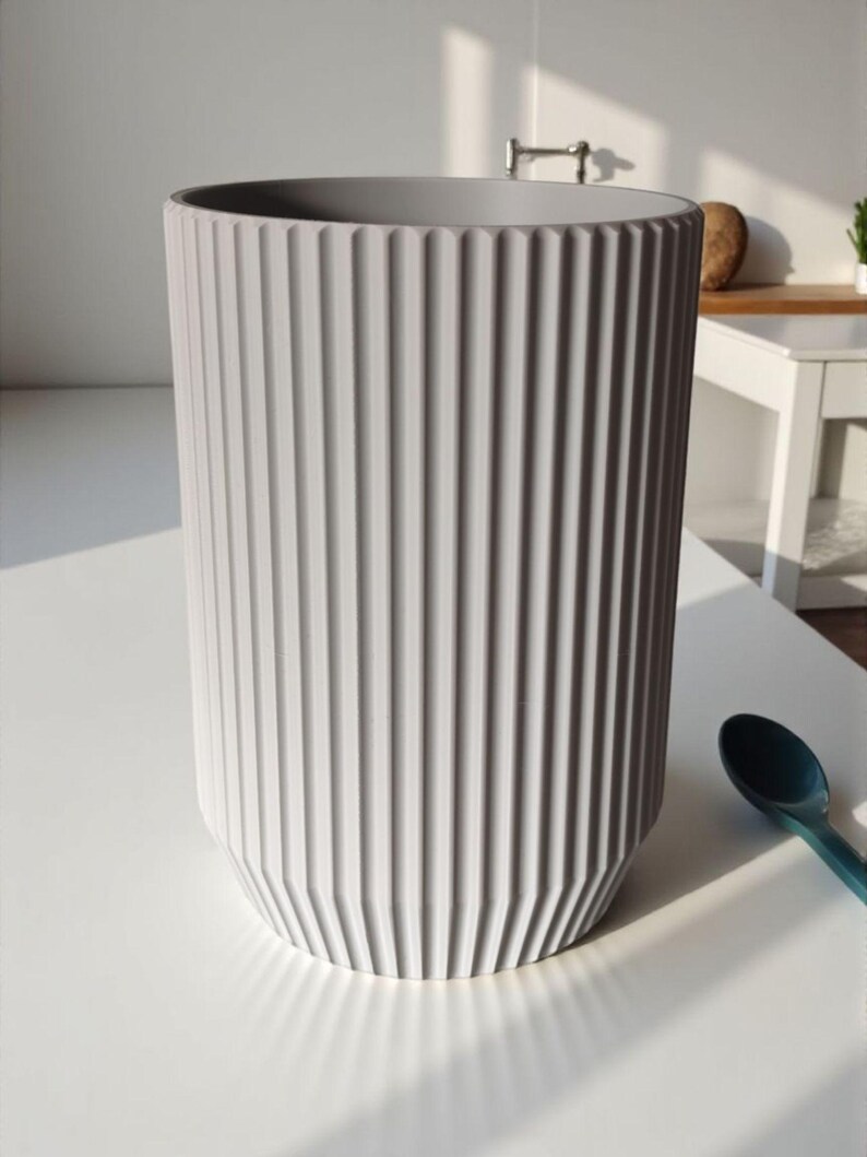 May include: A tall, gray cylindrical vase with vertical ridged detailing. The vase has a slightly tapered base and a smooth, open top. A teal spoon rests on the white surface next to the vase.