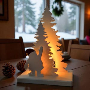 May include: A white Christmas-themed table decoration featuring a silhouette of a Santa Claus figure waving, with a backdrop of two illuminated Christmas trees. Two pine cones sit on the table. The decoration is on a white base.