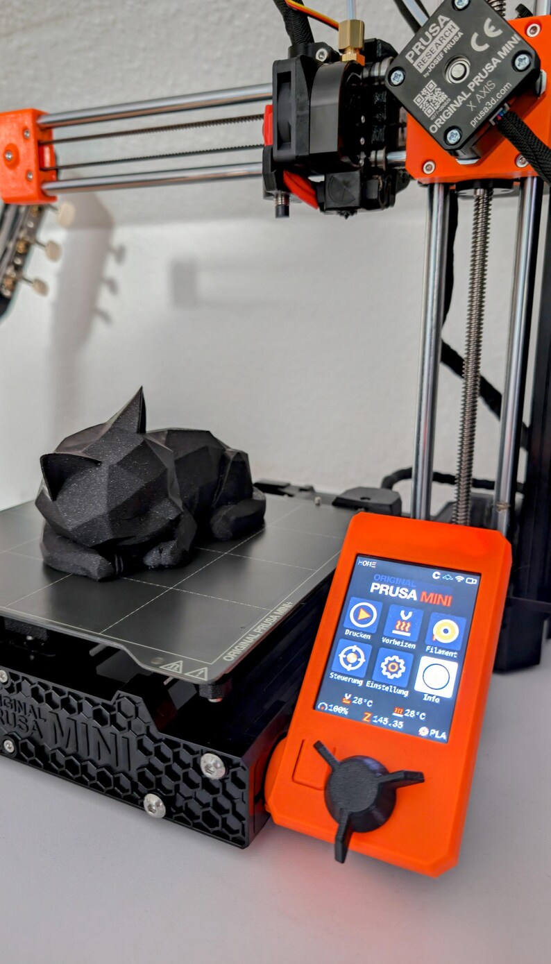 May include: A black 3D-printed cat figurine rests on the print bed of an orange and black Prusa Mini 3D printer. The printer's control panel displays settings. The printer is a desktop model.