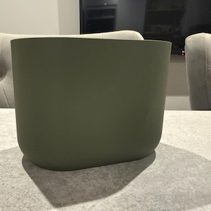 May include: An olive green, rectangular planter with rounded corners. The planter is sitting on a light gray surface. The planter's shadow is visible on the surface. The background includes two upholstered chairs and a television.