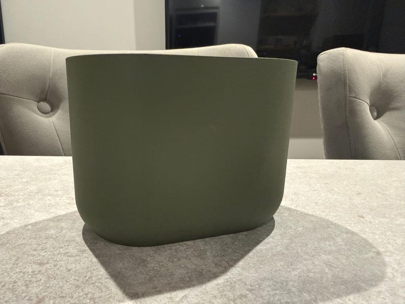 May include: An olive green, rectangular planter with rounded corners. The planter is sitting on a light gray surface. The planter's shadow is visible on the surface. The background includes two upholstered chairs and a television.