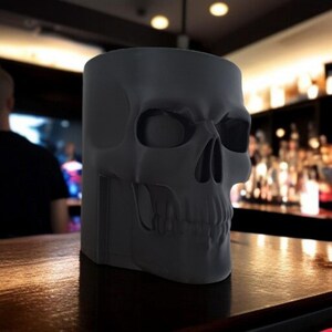May include: A matte black skull-shaped container, possibly a mug or planter, sits on a wooden surface. The container's design features detailed facial features, including eye sockets, a nose cavity, and teeth. The background is blurred, suggesting a bar setting.
