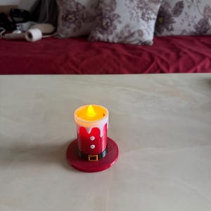 May include: A battery-operated candle with a Santa Claus design. The candle has a red base and body with white trim, a black belt, and a flickering yellow light. The candle sits on a red circular base.