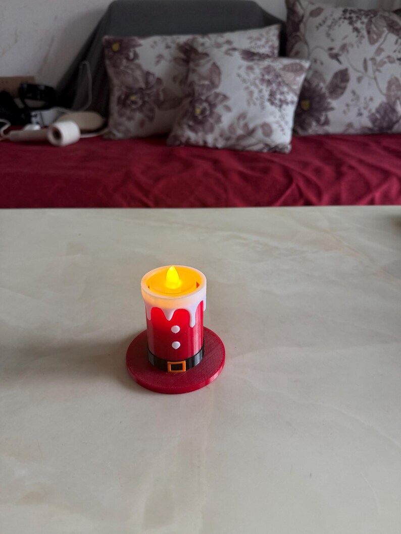May include: A battery-operated candle with a Santa Claus design. The candle has a red base and body with white trim, a black belt, and a flickering yellow light. The candle sits on a red circular base.