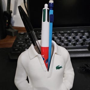 Puede incluir: Un portal&aacute;pices blanco con forma de polo, con un logotipo de cocodrilo verde en el pecho. Contiene un bol&iacute;grafo negro, un bol&iacute;grafo multicolor y un l&aacute;piz azul. Accesorio de escritorio.