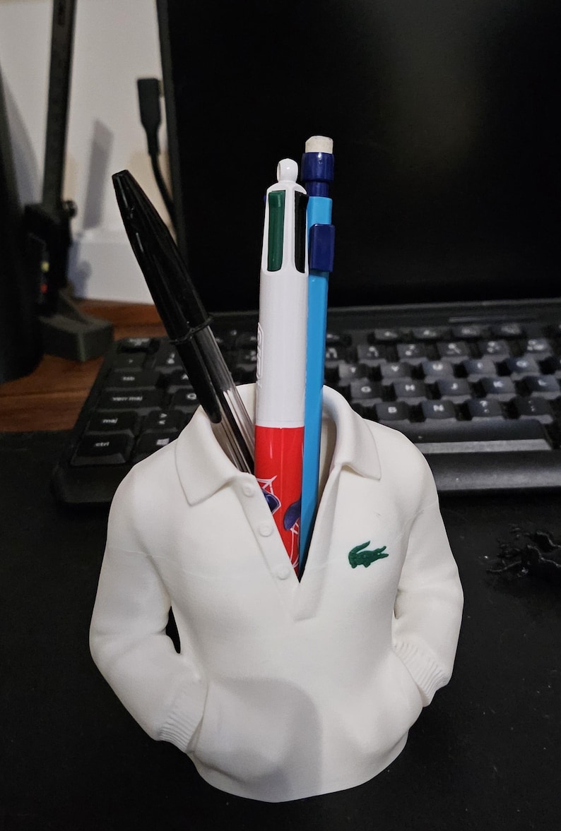 Puede incluir: Un portal&aacute;pices blanco con forma de polo, con un logotipo de cocodrilo verde en el pecho. Contiene un bol&iacute;grafo negro, un bol&iacute;grafo multicolor y un l&aacute;piz azul. Accesorio de escritorio.