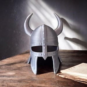 May include: A silver-colored Viking helmet with curved horns, resting on a wooden surface. The helmet has eye and mouth openings and is next to an open book. The background is a dark gray wall.