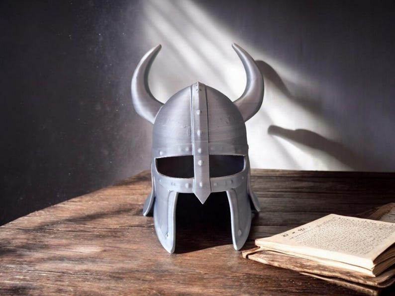 May include: A silver-colored Viking helmet with curved horns, resting on a wooden surface. The helmet has eye and mouth openings and is next to an open book. The background is a dark gray wall.