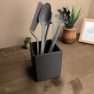 May include: A set of gray kitchen utensils, including a spatula, spoon, slotted spoon, and tongs, displayed in a black square holder. The utensils are made of a matte material and are arranged on a wooden surface. A small potted plant and a framed picture are in the background.