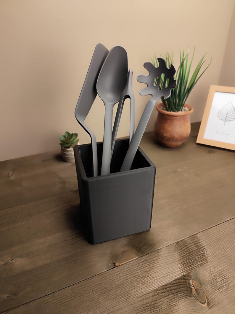 May include: A set of gray kitchen utensils, including a spatula, spoon, slotted spoon, and tongs, displayed in a black square holder. The utensils are made of a matte material and are arranged on a wooden surface. A small potted plant and a framed picture are in the background.