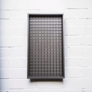 Op de afbeelding: Een rechthoekige, donkergrijze tray met een rasterpatroon van kleine vierkantjes. De tray heeft een opstaande rand en staat tegen een witte bakstenen muur. Het oppervlak van de tray heeft een textuur.