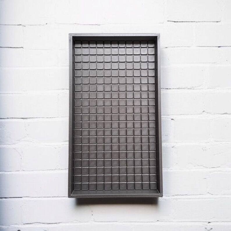 Op de afbeelding: Een rechthoekige, donkergrijze tray met een rasterpatroon van kleine vierkantjes. De tray heeft een opstaande rand en staat tegen een witte bakstenen muur. Het oppervlak van de tray heeft een textuur.