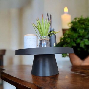 May include: A black cake stand displays a small potted plant, a candle, and a reed diffuser. The stand is on a wooden table. The items on the stand are for home decor.
