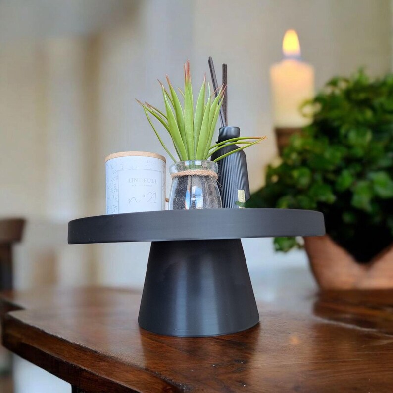 May include: A black cake stand displays a small potted plant, a candle, and a reed diffuser. The stand is on a wooden table. The items on the stand are for home decor.