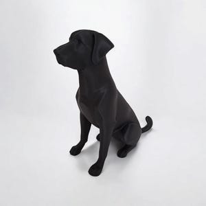 May include: A solid black Labrador Retriever statue in a seated position. The dog is facing left, with its head turned slightly upward. The statue has a smooth, matte finish and is set against a white background.