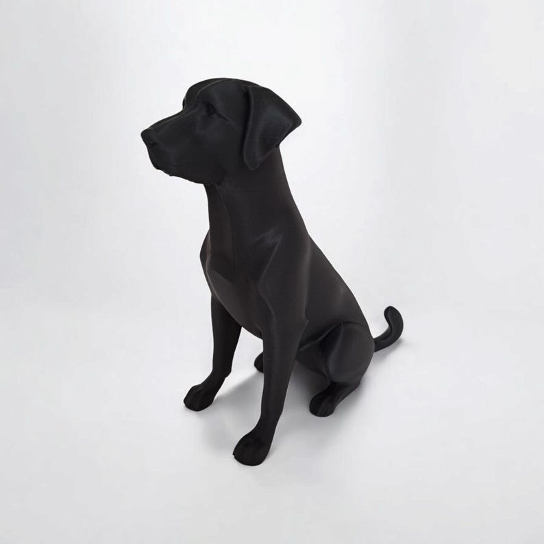 May include: A solid black Labrador Retriever statue in a seated position. The dog is facing left, with its head turned slightly upward. The statue has a smooth, matte finish and is set against a white background.