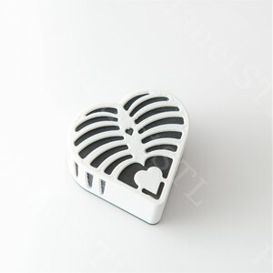 May include: A white, heart-shaped decorative object with a black interior. The object has a lattice design on the top and a small heart cutout on the bottom. The object is on a white surface.