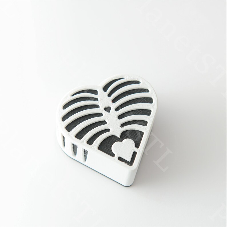 May include: A white, heart-shaped decorative object with a black interior. The object has a lattice design on the top and a small heart cutout on the bottom. The object is on a white surface.