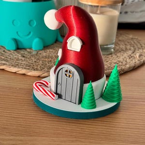 以下が含まれることがあります： サンタ帽の形をした、気まぐれなクリスマスをテーマにした装飾品。灰色のドア、緑の木、キャンディケインが特徴です。帽子は赤い色で、白いポンポンが付いており、ベースはティール色です。