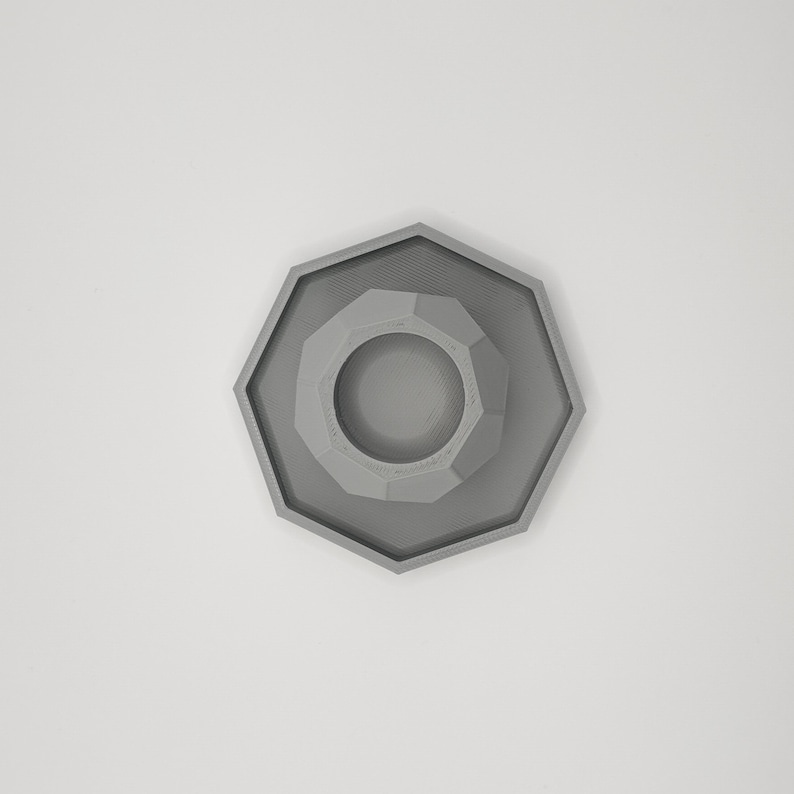 May include: A gray, geometric, octagonal object with a central circular depression. The object has a layered design, with each layer slightly smaller than the one below. The overall shape is symmetrical and modern.