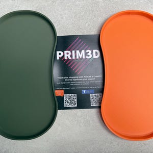 May include: Two oval trays, one in olive green and the other in bright orange, are shown. The trays have a smooth, curved design. A card with the text "PRIM3D" and other text is placed between the trays.