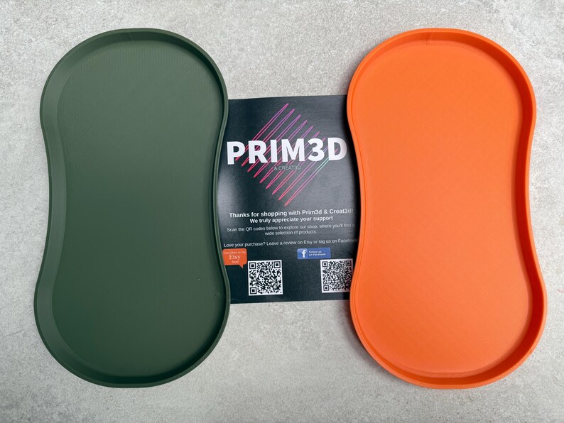 May include: Two oval trays, one in olive green and the other in bright orange, are shown. The trays have a smooth, curved design. A card with the text "PRIM3D" and other text is placed between the trays.