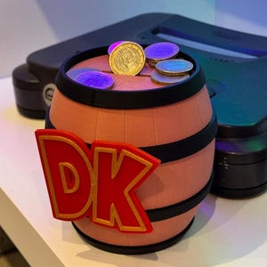 May include: A novelty money bank shaped like a barrel, featuring the letters "DK" in red with gold trim. The barrel is light brown with black bands and contains coins. A black Nintendo 64 console is in the background.