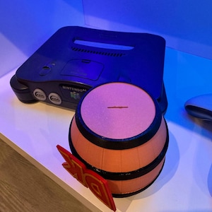 May include: A black Nintendo 64 game console with a barrel-shaped object and a red and gold DK logo. The barrel is orange with black bands. The DK logo is red with gold accents. The console is on a white surface.
